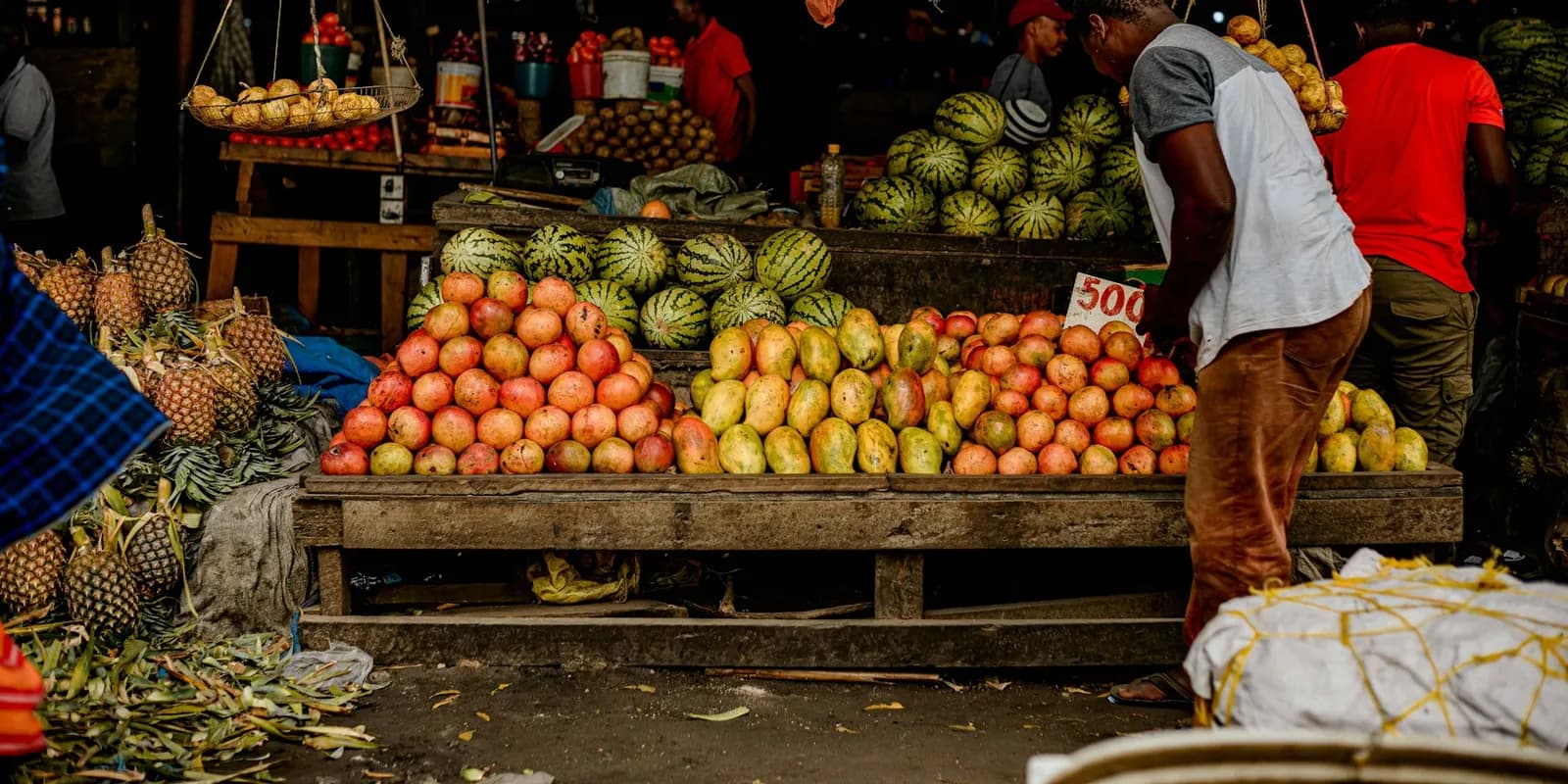 A trader arranging mangoes and watermelons at a busy fruit market